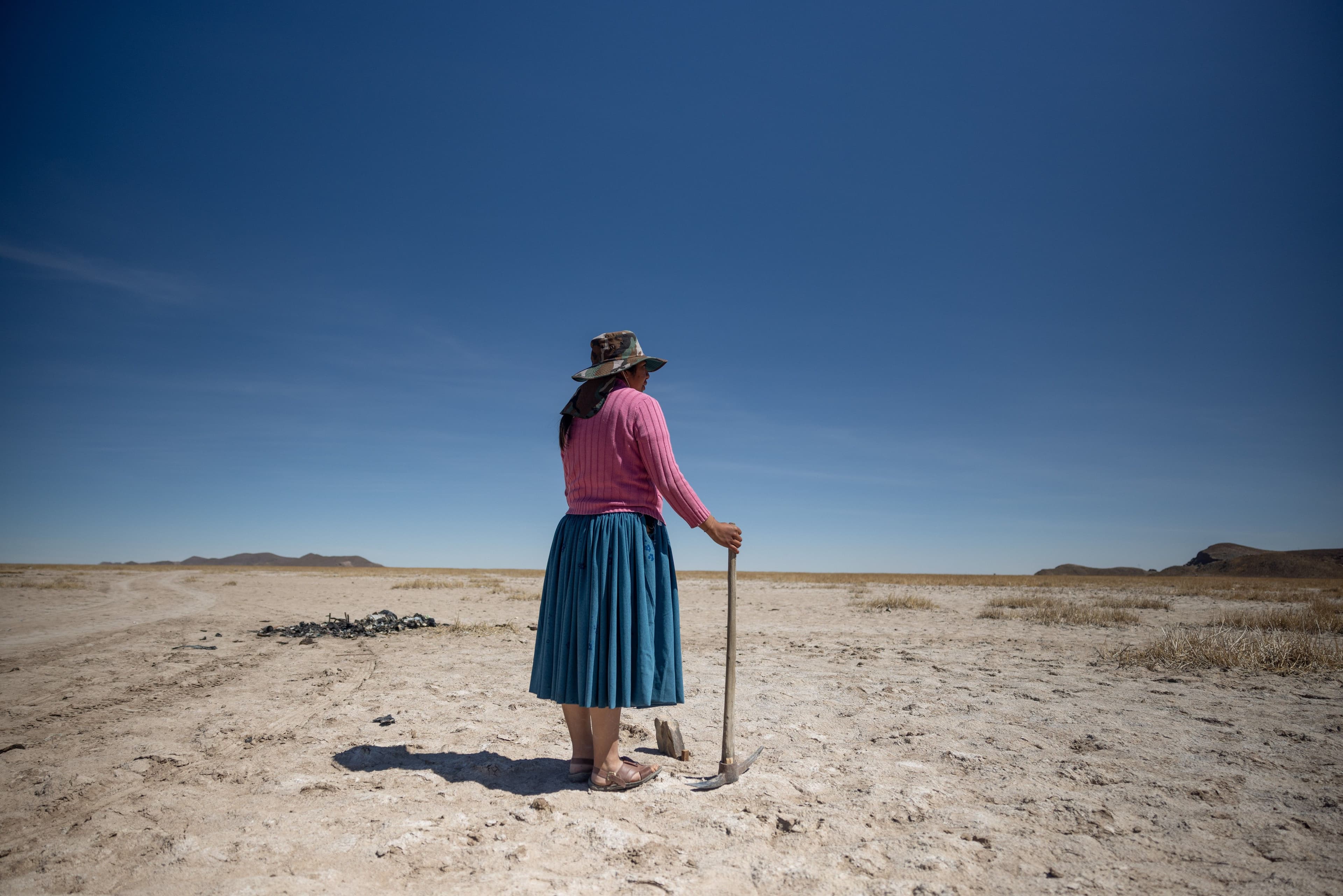 Seen from behind, a woman in a pink sweater, blue skirt, and wide-brimmed hat stands alone on a vast, parched plain, leaning on a farming tool. The expansive blue sky and barren landscape give the image a powerful sense of solitude and environmental hardship.