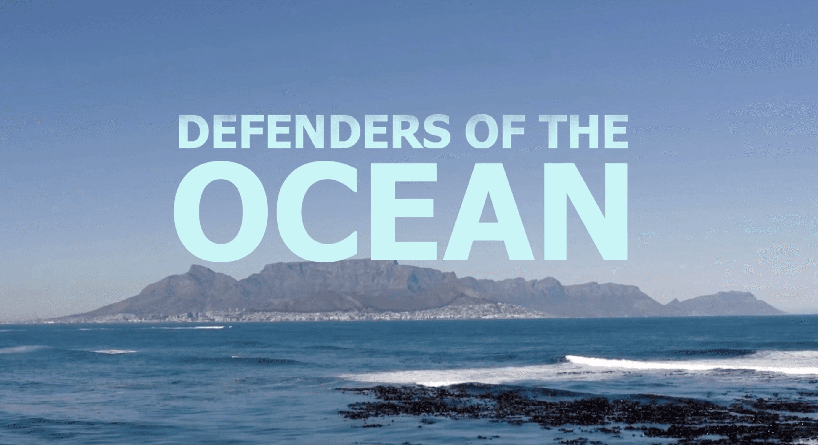 A sweeping coastal view of Cape Town's Table Mountain rising above the city, framed by the vivid blue Atlantic Ocean and rocky shoreline in the foreground. The wide, sun-drenched landscape carries a sense of both grandeur and vulnerability.