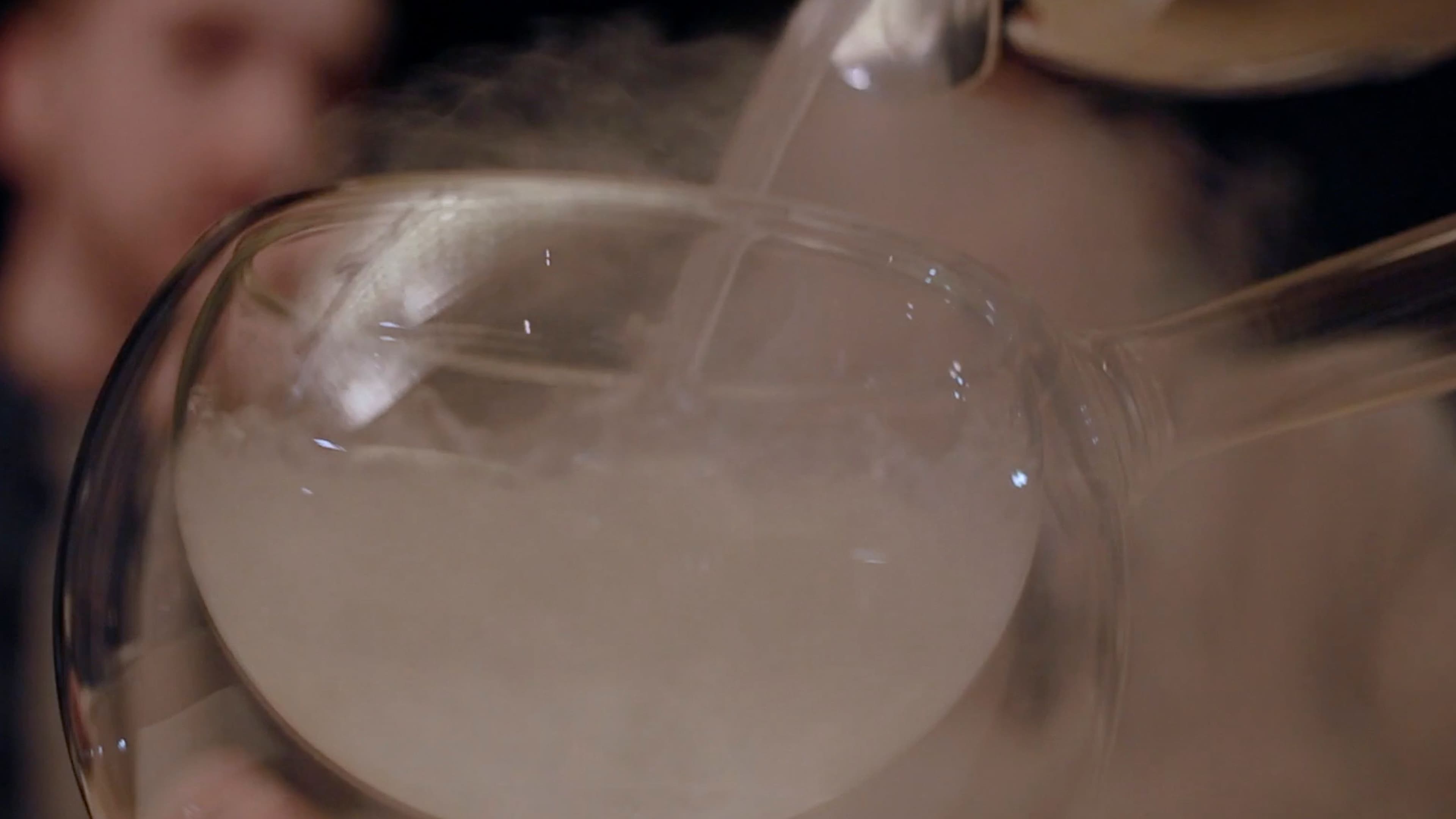 A theatrical tableside pour into a wide glass releases a cloud of mist, lit by warm candlelight in the background.