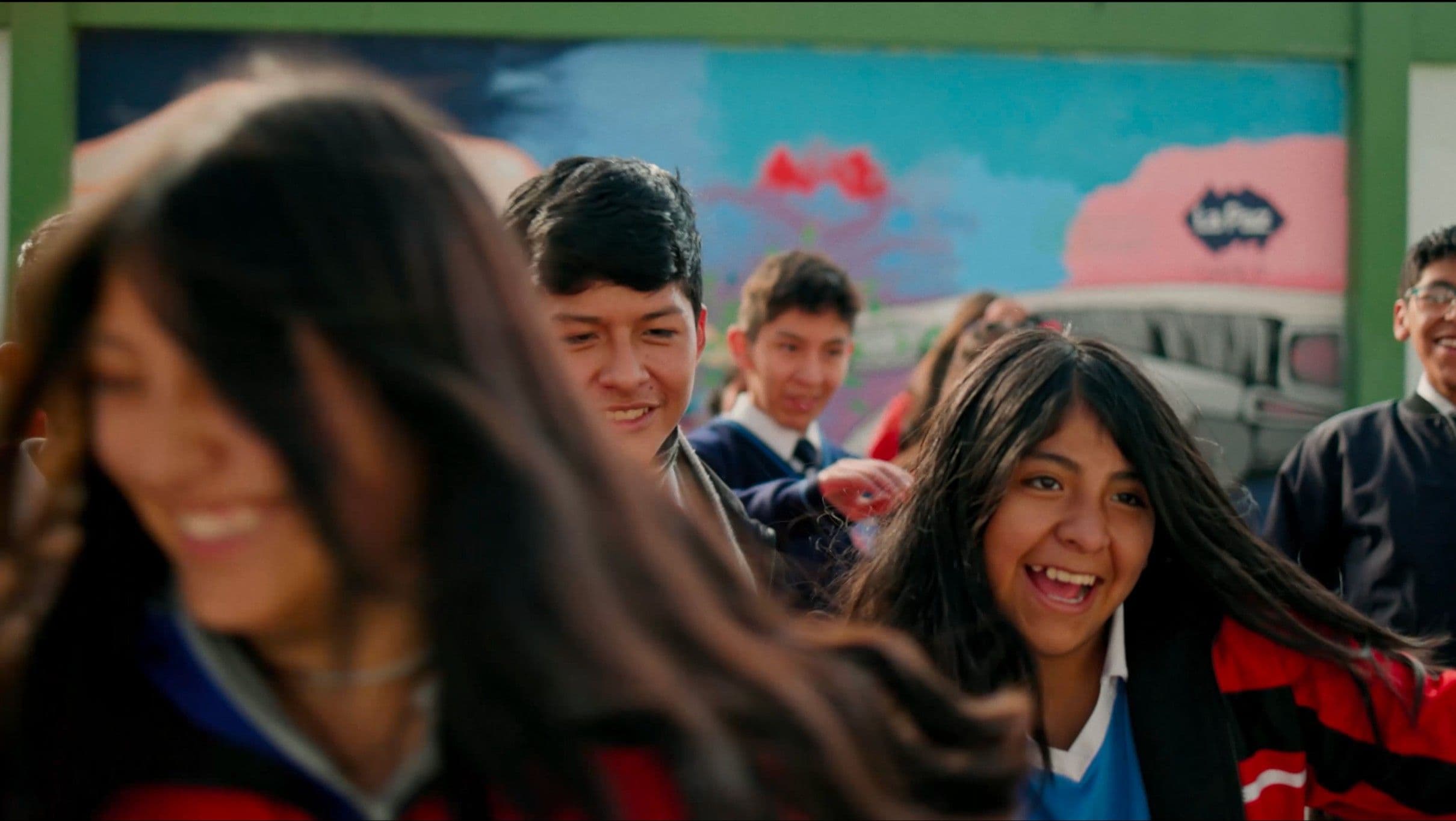 A group of laughing Latin American teenagers in school uniforms move energetically in front of a vibrant painted mural. The dynamic, motion-filled shot captures the joy and energy of youth.