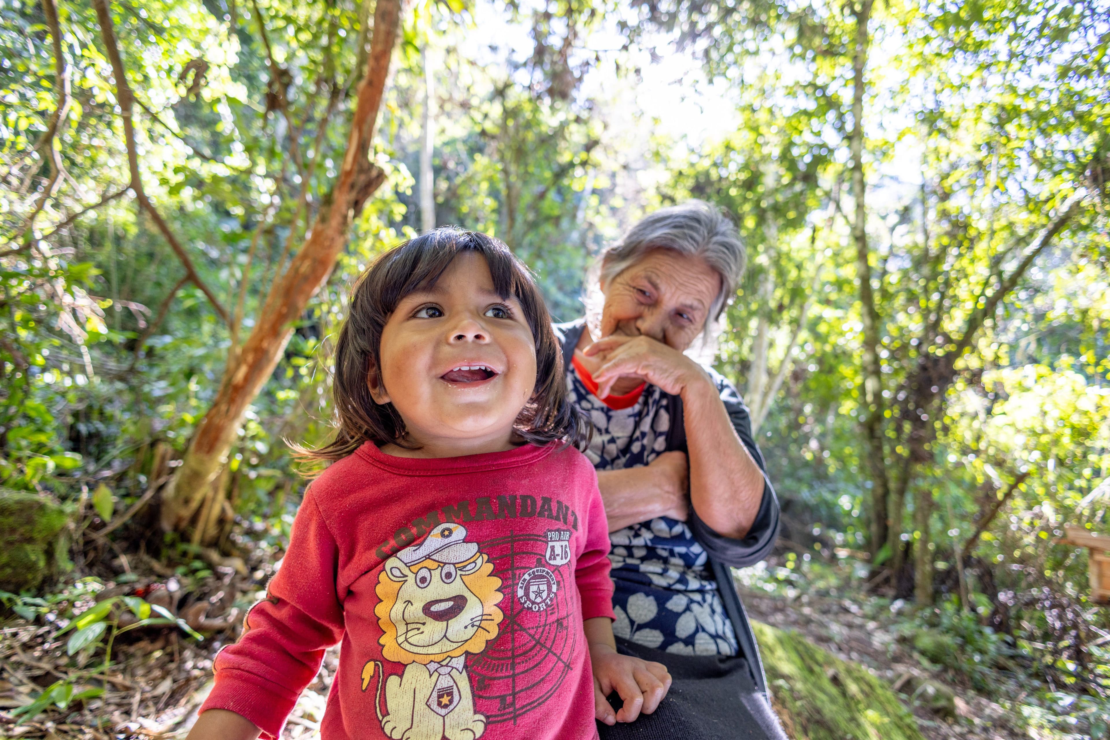A young child in a red lion-print sweatshirt gazes upward with delight in a sun-dappled forest, while an older woman laughs warmly behind them. The low-angle, close-up framing captures an intimate moment of intergenerational joy in nature.