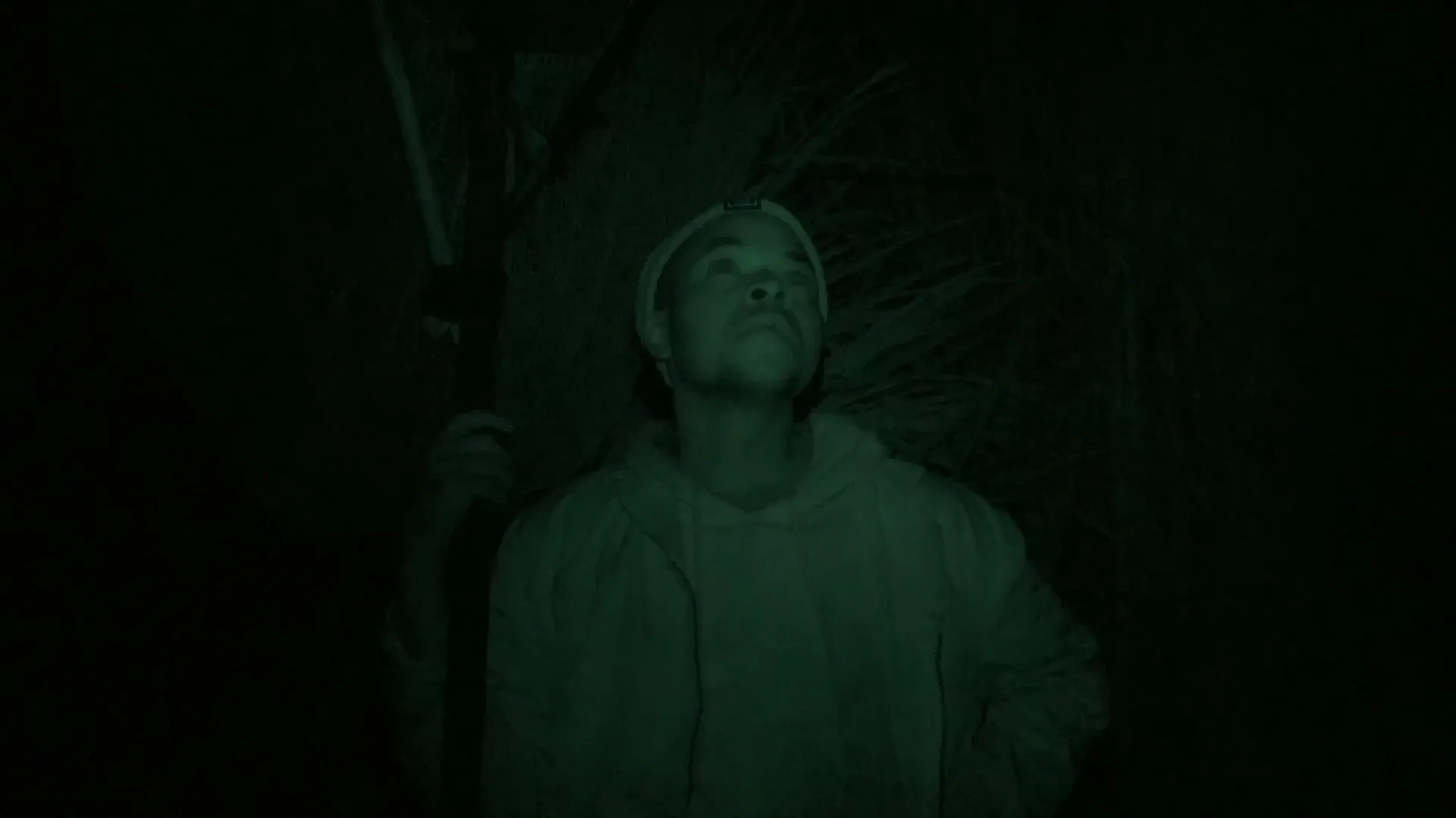 A night-vision shot of a person standing outdoors after dark, looking upward while holding a tool.