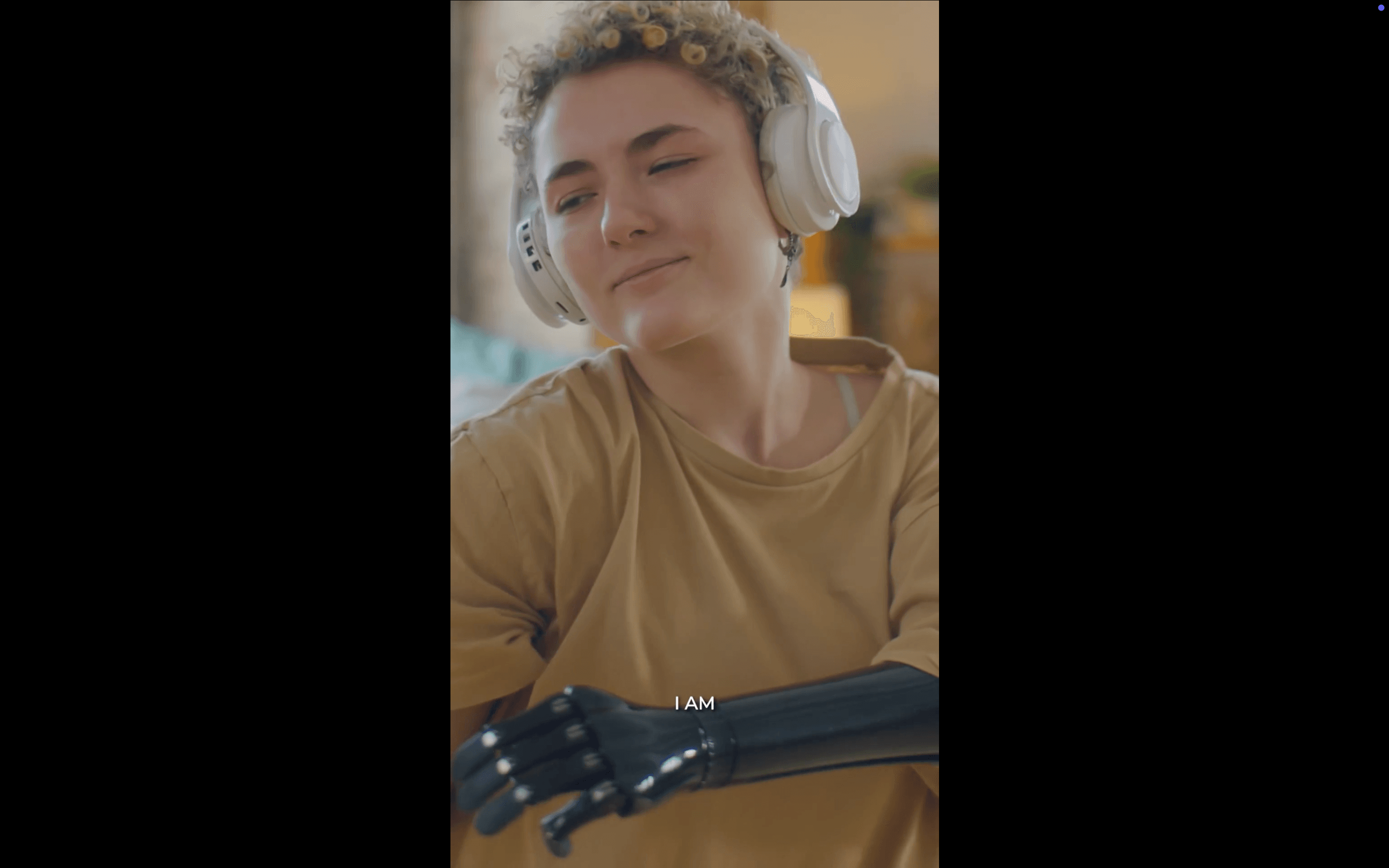 A young person with short curly hair and wireless headphones sits with a relaxed smile, a dark prosthetic arm resting in the foreground, with the subtitle "I AM." The warm, intimate lighting and quiet confidence make the shot quietly powerful.