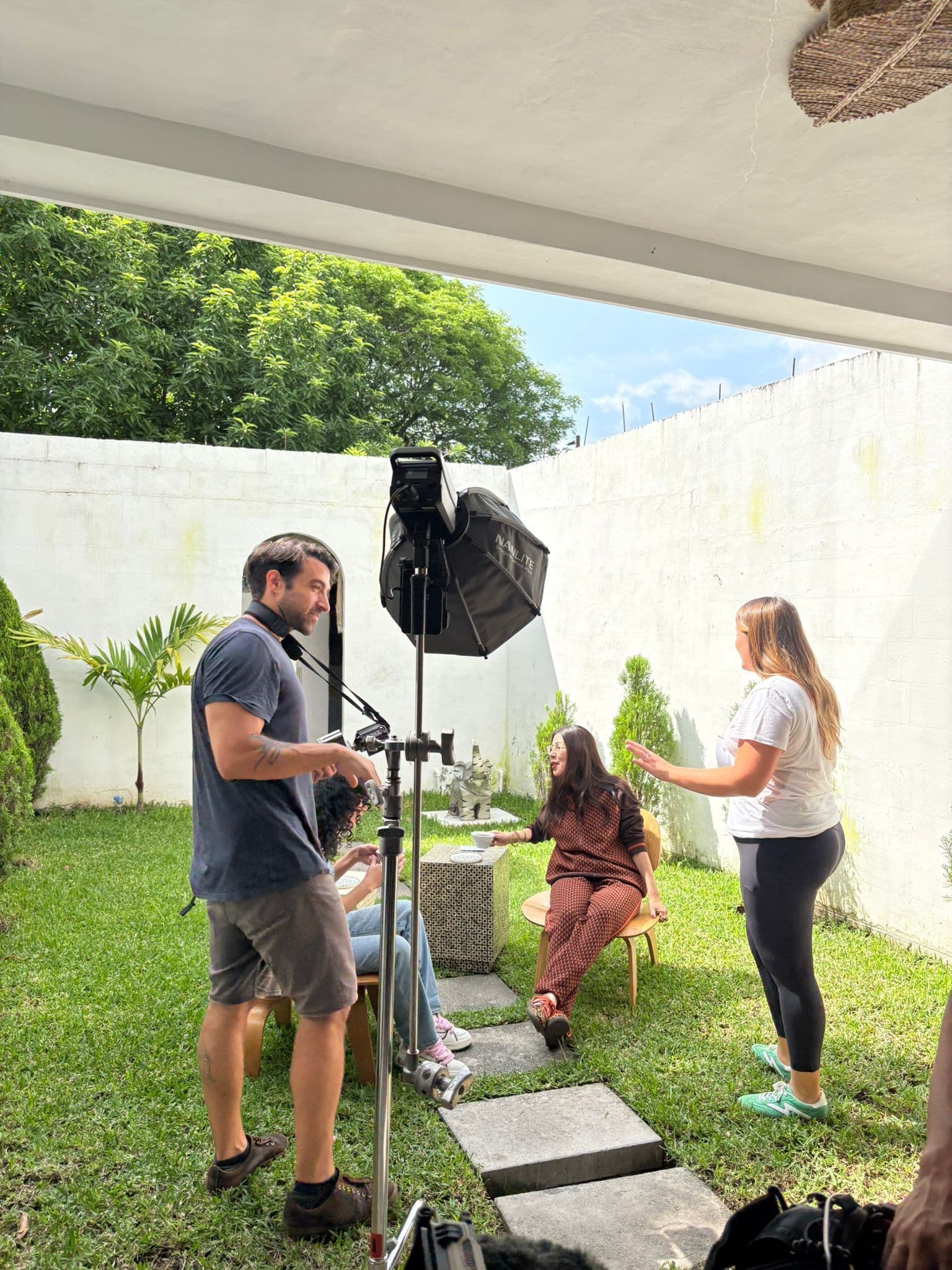 A film crew sets up a boom mic and light rig to film an interview in a lush garden courtyard, two women conversing while seated.