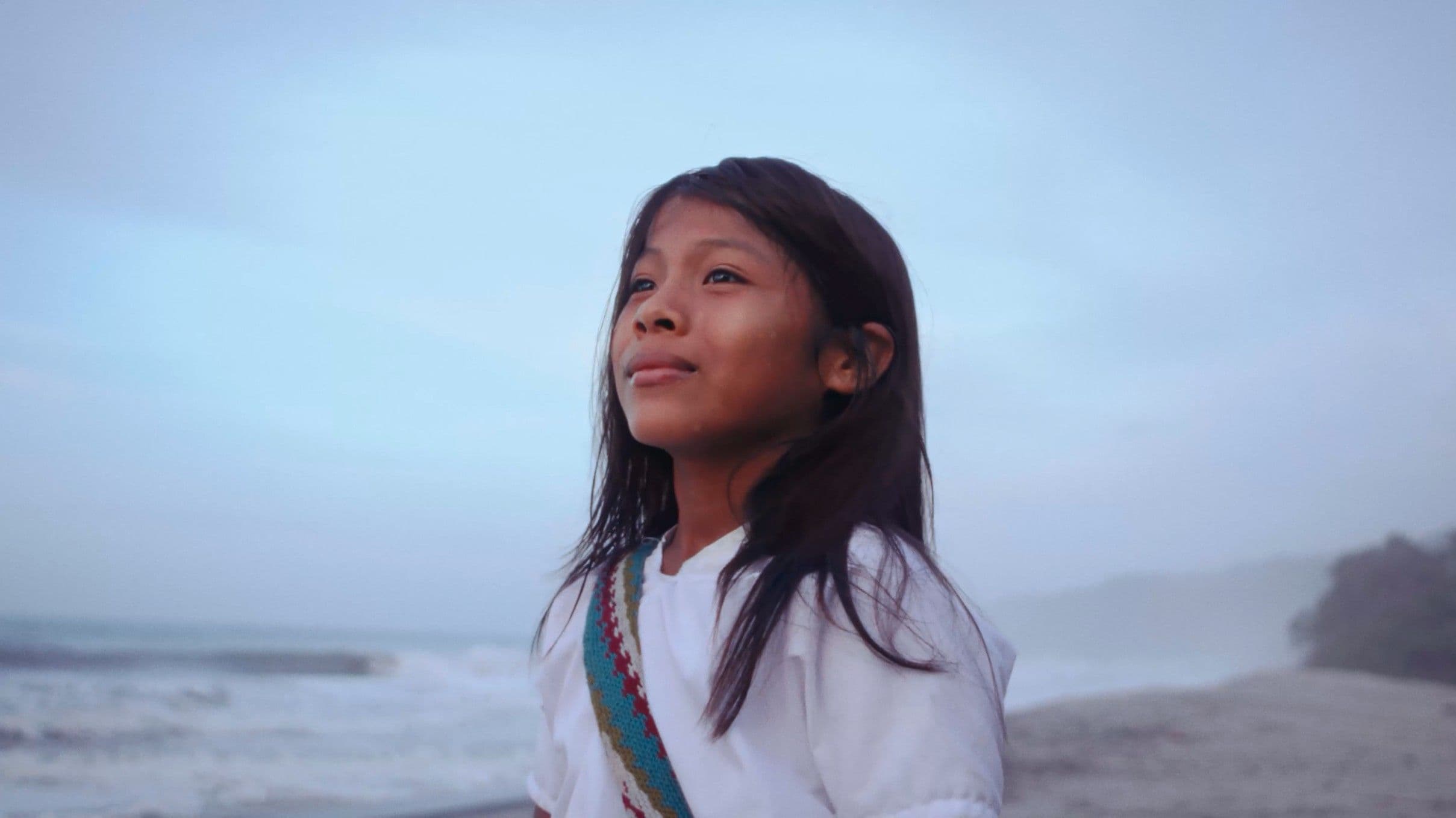 A young Latin American girl with long dark hair and a woven sash looks upward with quiet wonder, standing before a misty coastal horizon. The soft blue tones and open sky give the shot a serene, cinematic feel.