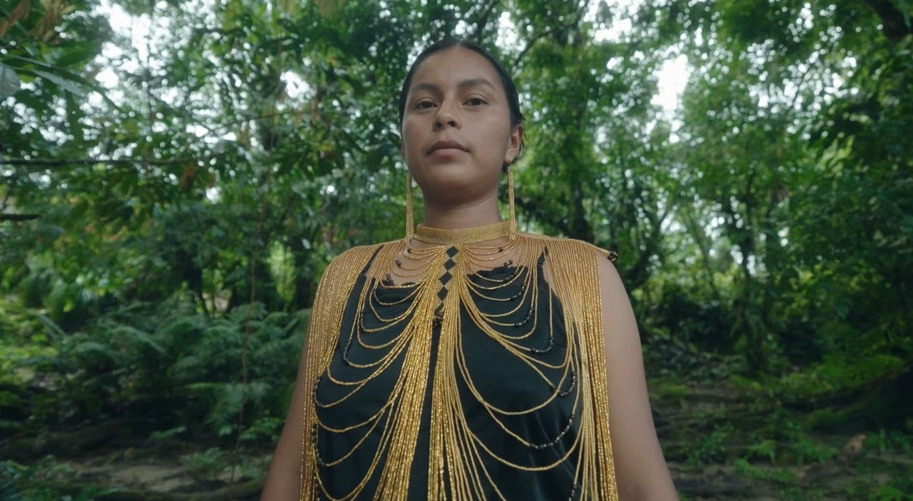 A young indigenous Latin American woman stands in a lush green forest, wearing an elaborate golden beaded collar and matching earrings over a dark garment. Her direct, steady gaze and striking adornment convey quiet strength and cultural identity.