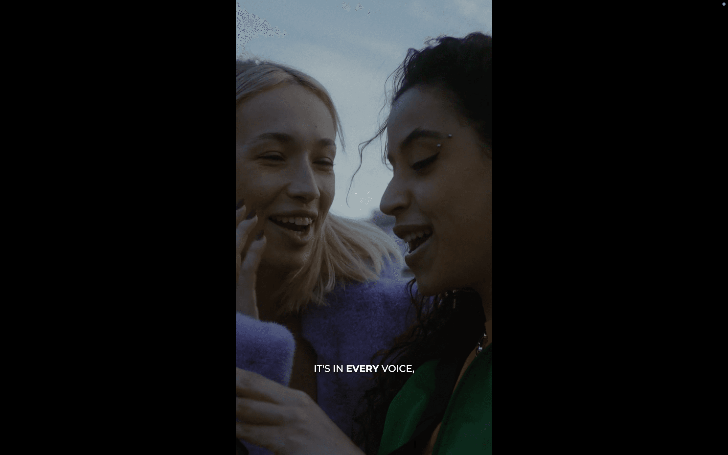 Two young women — one blonde, one dark-haired — lean close together laughing, with the subtitle "IT'S IN EVERY VOICE," against a muted outdoor backdrop. The intimate, candid framing conveys warmth, connection, and shared joy.
