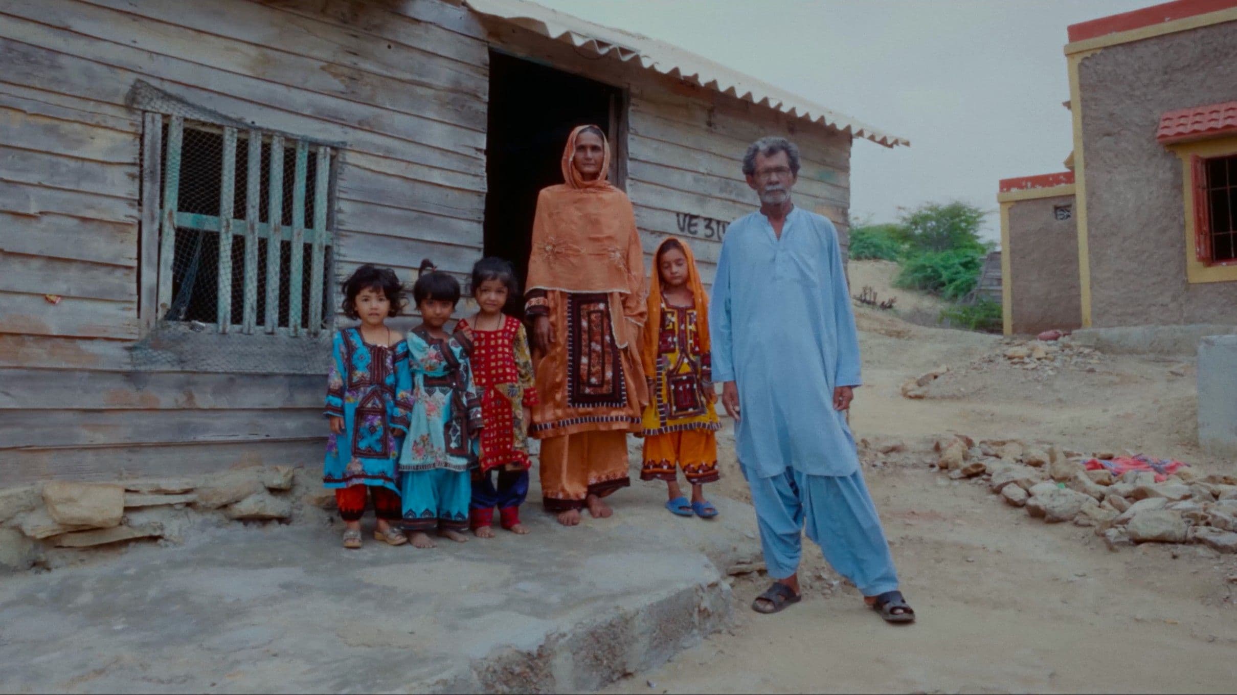 A South Asian family — an older man in a light blue shalwar kameez, a woman in an orange dupatta, and four young children in vibrant traditional dress — stand together outside a modest wooden home in a rural village. The documentary-style still captures the quiet dignity of everyday life.
