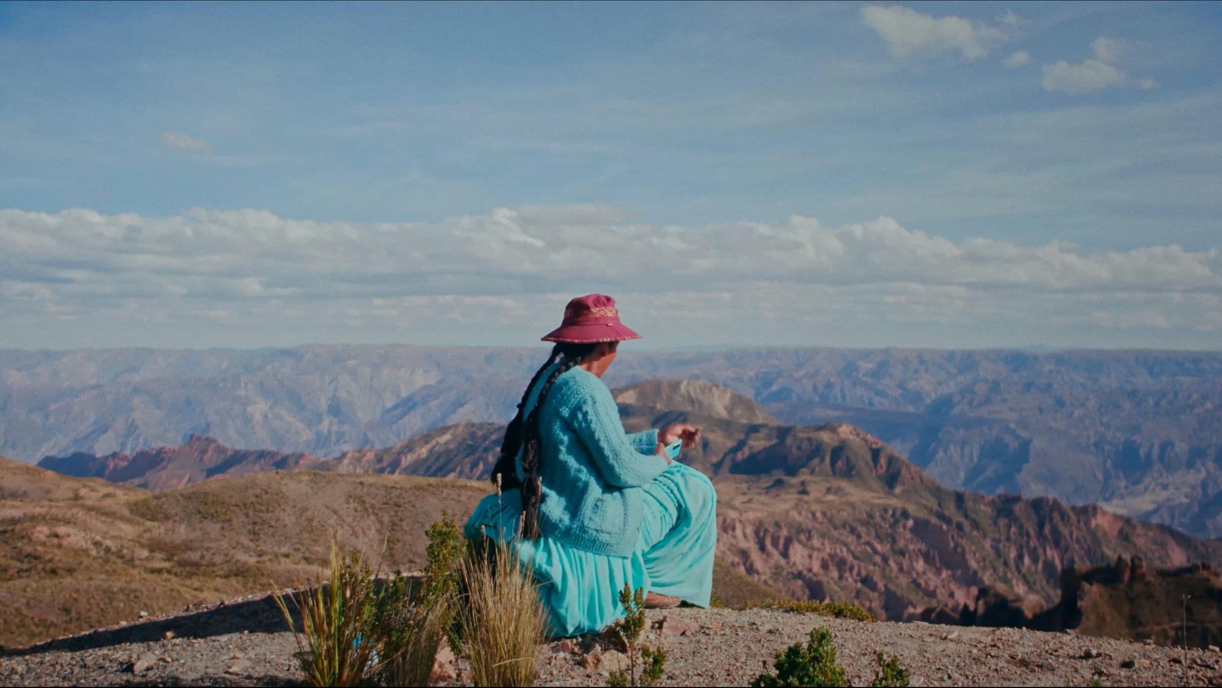 A woman in a bright turquoise dress and pink hat sits alone on a mountain ridge, gazing out over a sweeping Andean valley under a vast blue sky. The wide cinematic frame emphasises her solitude against the immensity of the landscape.