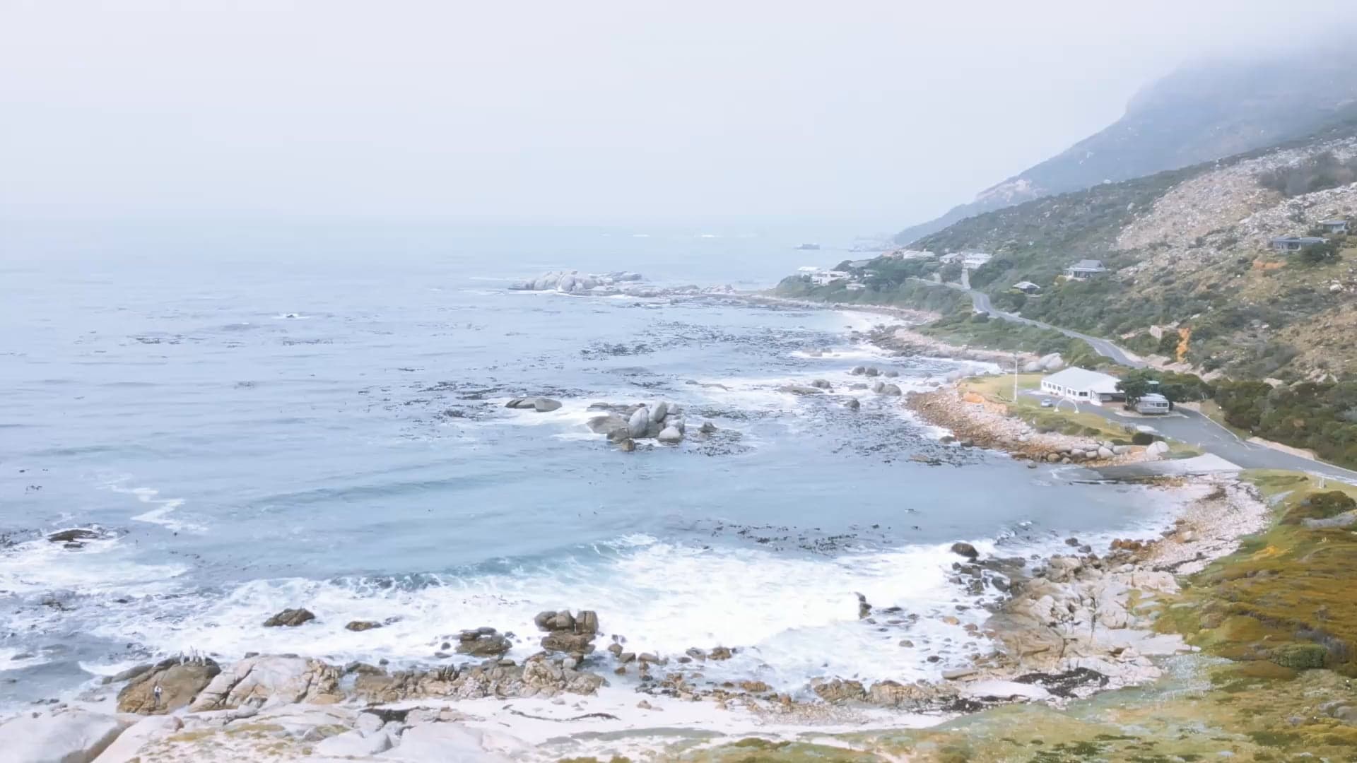 An aerial view of a rocky South African coastline with waves breaking near a small coastal settlement.