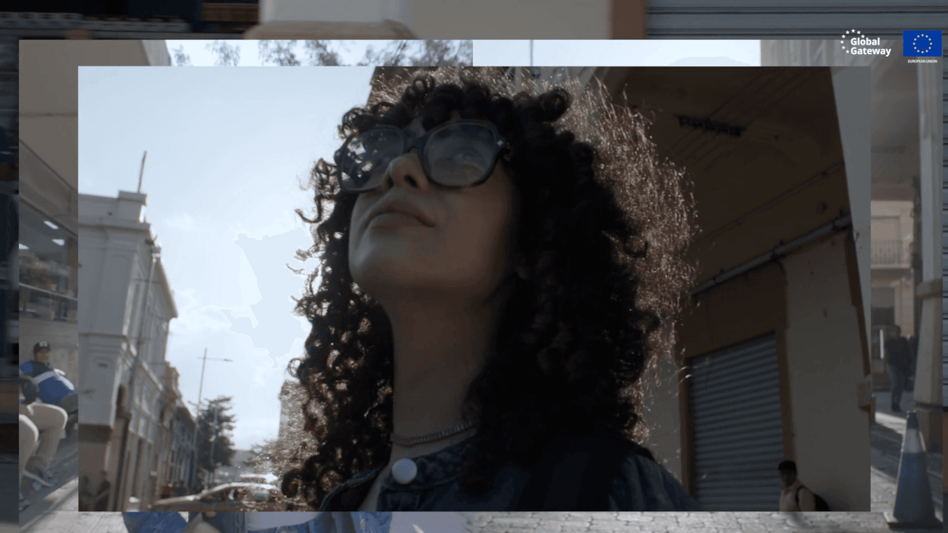 A young woman with curly hair and sunglasses tilts her face up toward the sun on a street in San Salvador.