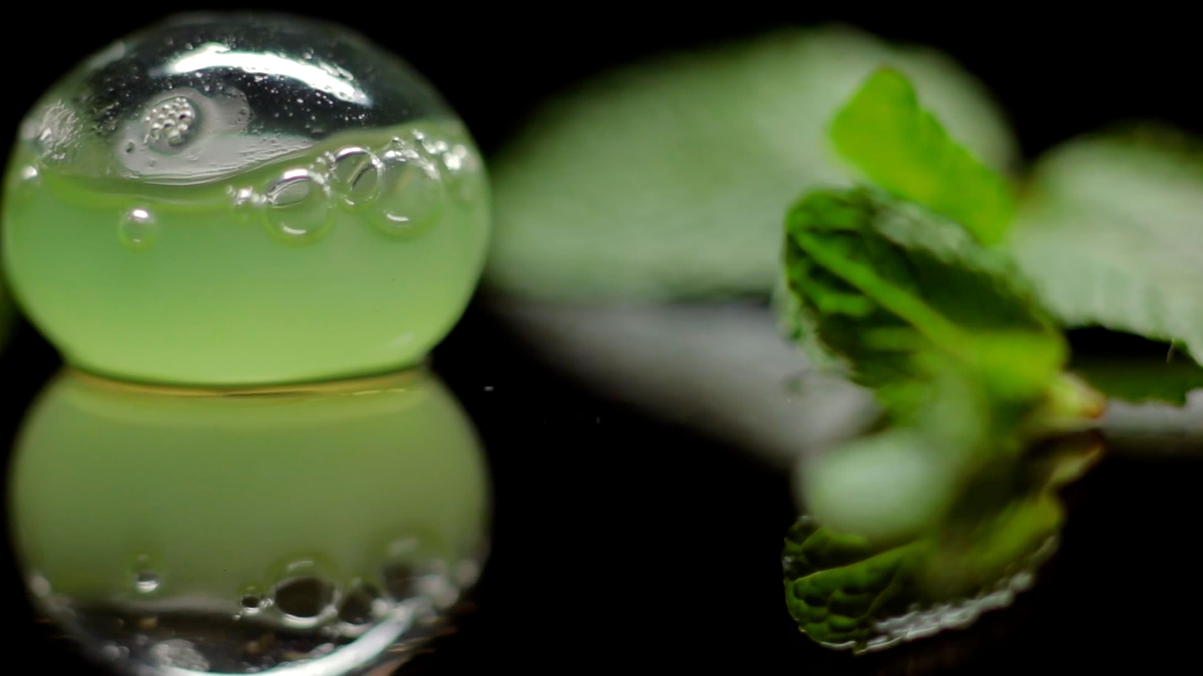 A translucent green spherified dish, a molecular gastronomy creation at minibar, sits on a mirrored black surface beside fresh mint leaves, its reflection glowing below.