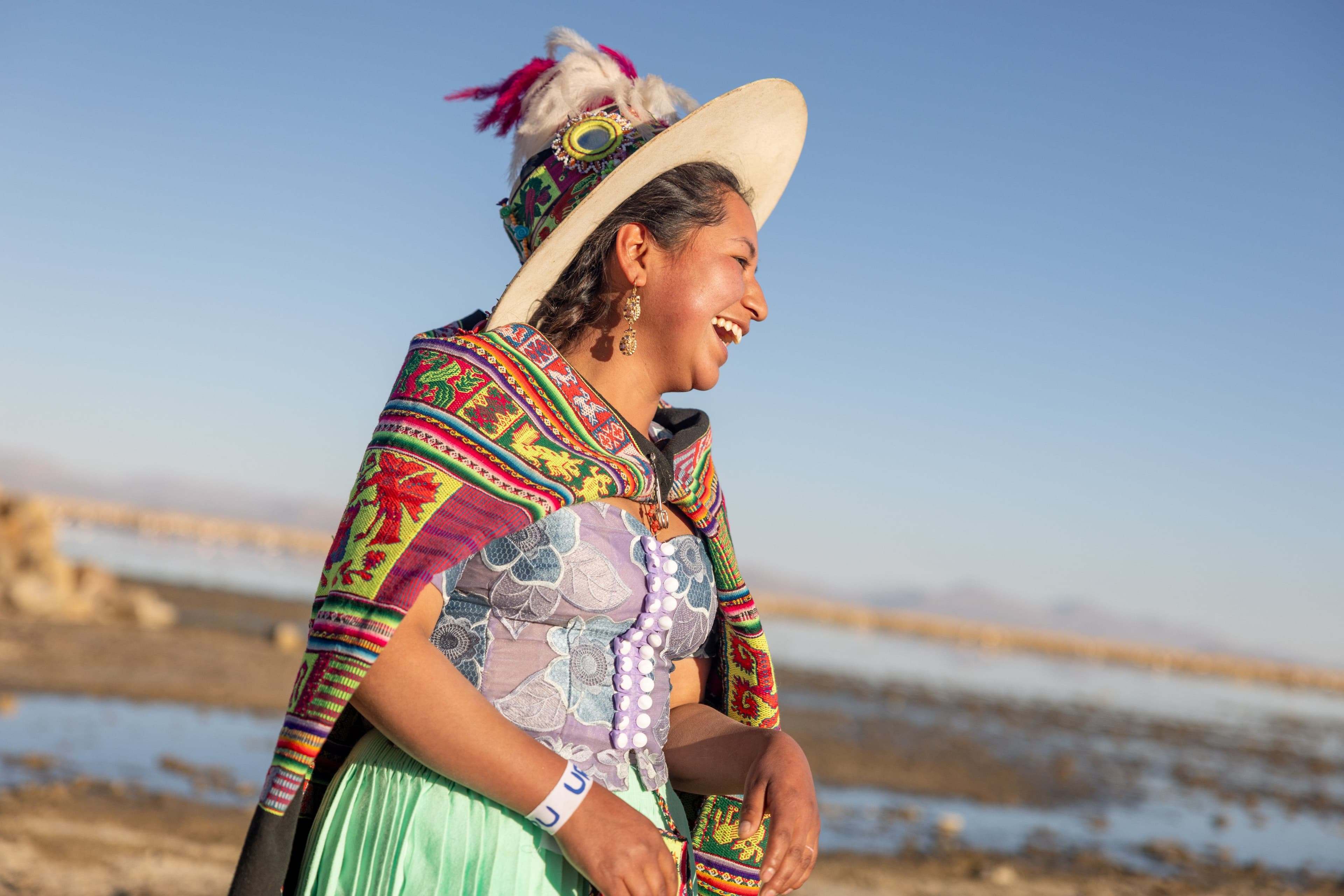A Bolivian woman in vibrant traditional dress — an embroidered shawl, lace bodice, green skirt and feathered hat — laughs joyfully in profile against a wide open sky and salt flat landscape. The warm golden light and candid expression make the image feel alive with cultural pride and spontaneous happiness.