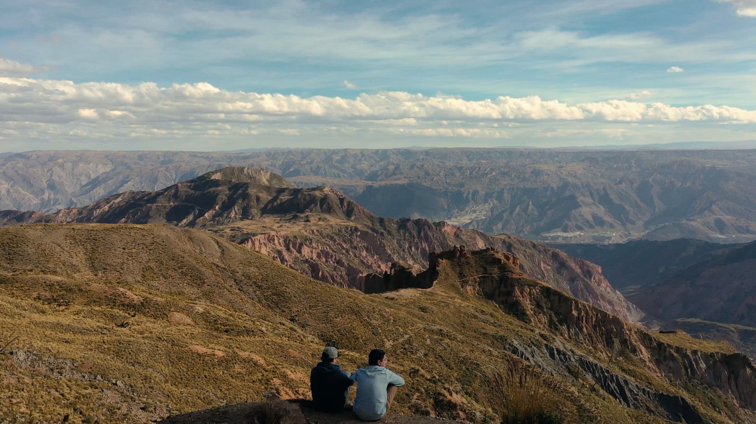 Two figures sit side by side on a mountain ridge, dwarfed by a vast, dramatic landscape of rugged Andean valleys stretching to the horizon. The wide shot conveys scale, solitude, and quiet connection.