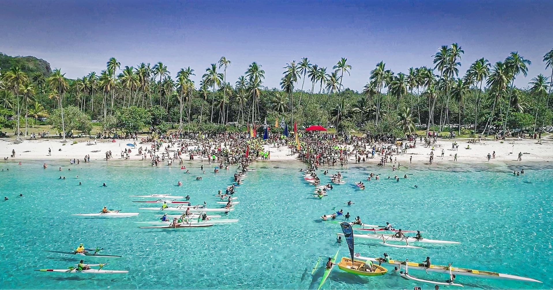 An aerial view of a palm-fringed beach packed with a community gathering, with dozens of outrigger canoes lined up in the turquoise lagoon.