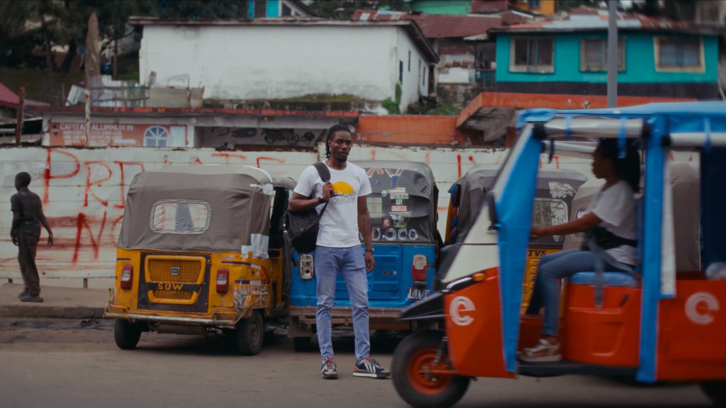A young Black man in a white t-shirt and jeans stands confidently between colourful tuk-tuks on a busy urban street in Monrovia, Liberia. The vivid street scene behind him captures the energy and texture of city life.