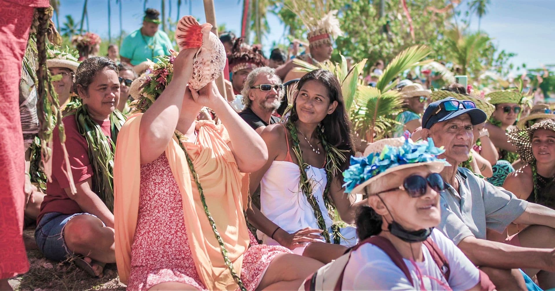 A seated crowd wearing floral leis and woven headdresses watches as a woman blows a conch shell at a community ceremony.