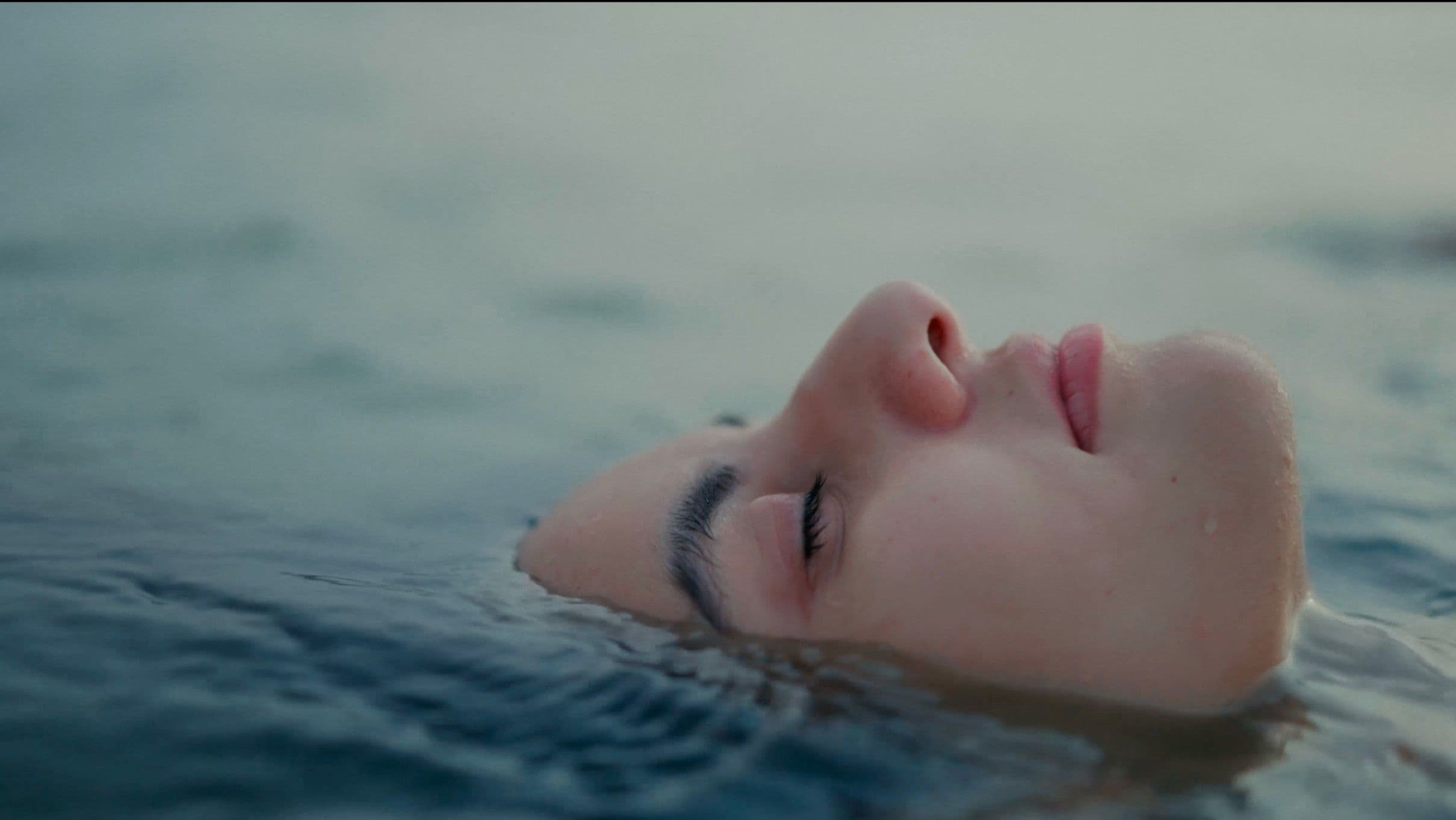 A woman's face floats just above the surface of still, grey-blue water, eyes closed in serene stillness. The extreme close-up and cool tones create a deeply atmospheric, meditative cinematic moment.