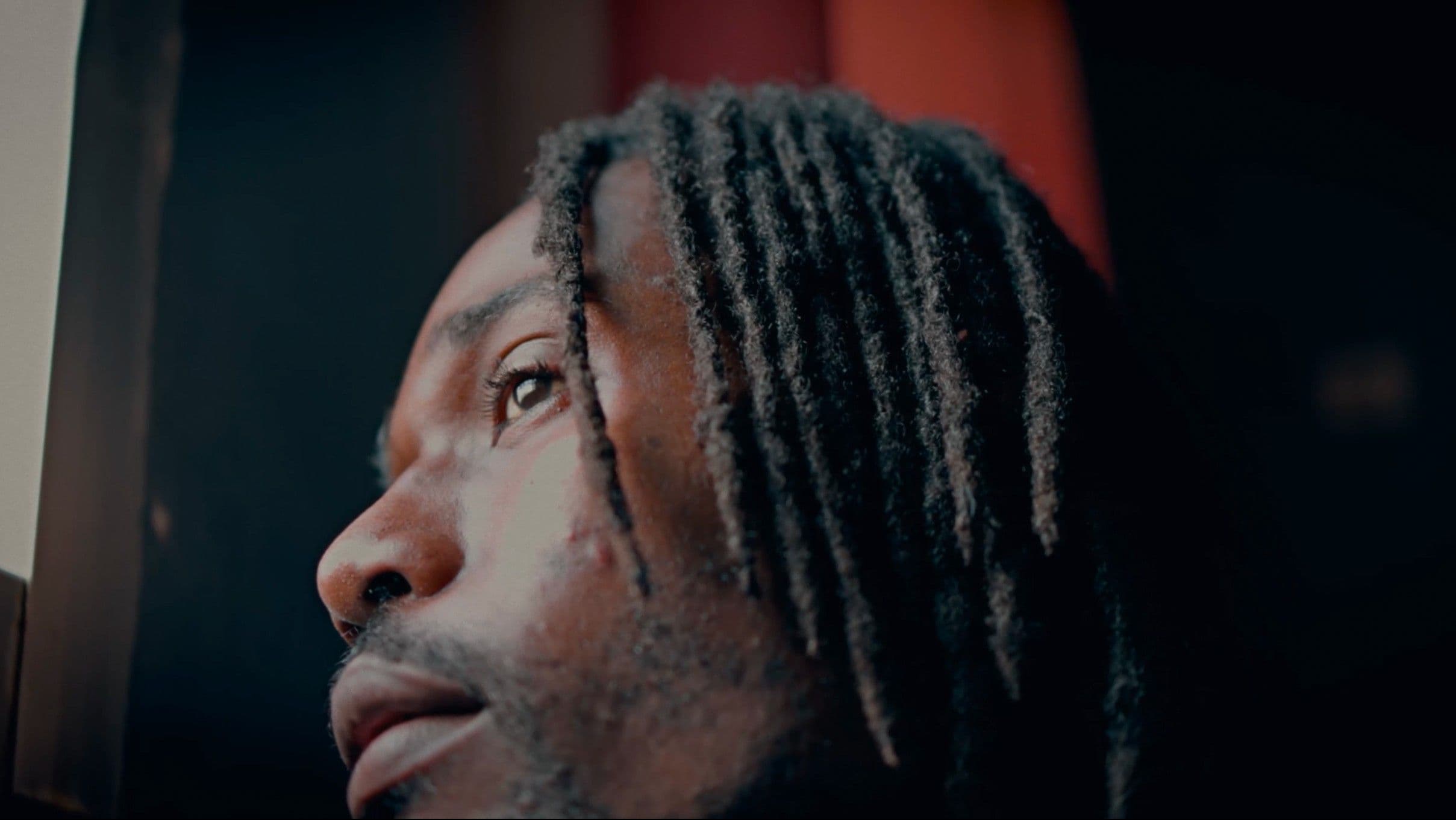 A tight close-up of a Black man with dreadlocks gazing upward, his face partially lit against a dark, shadowy background. The dramatic lighting gives the shot a deeply introspective, cinematic quality.