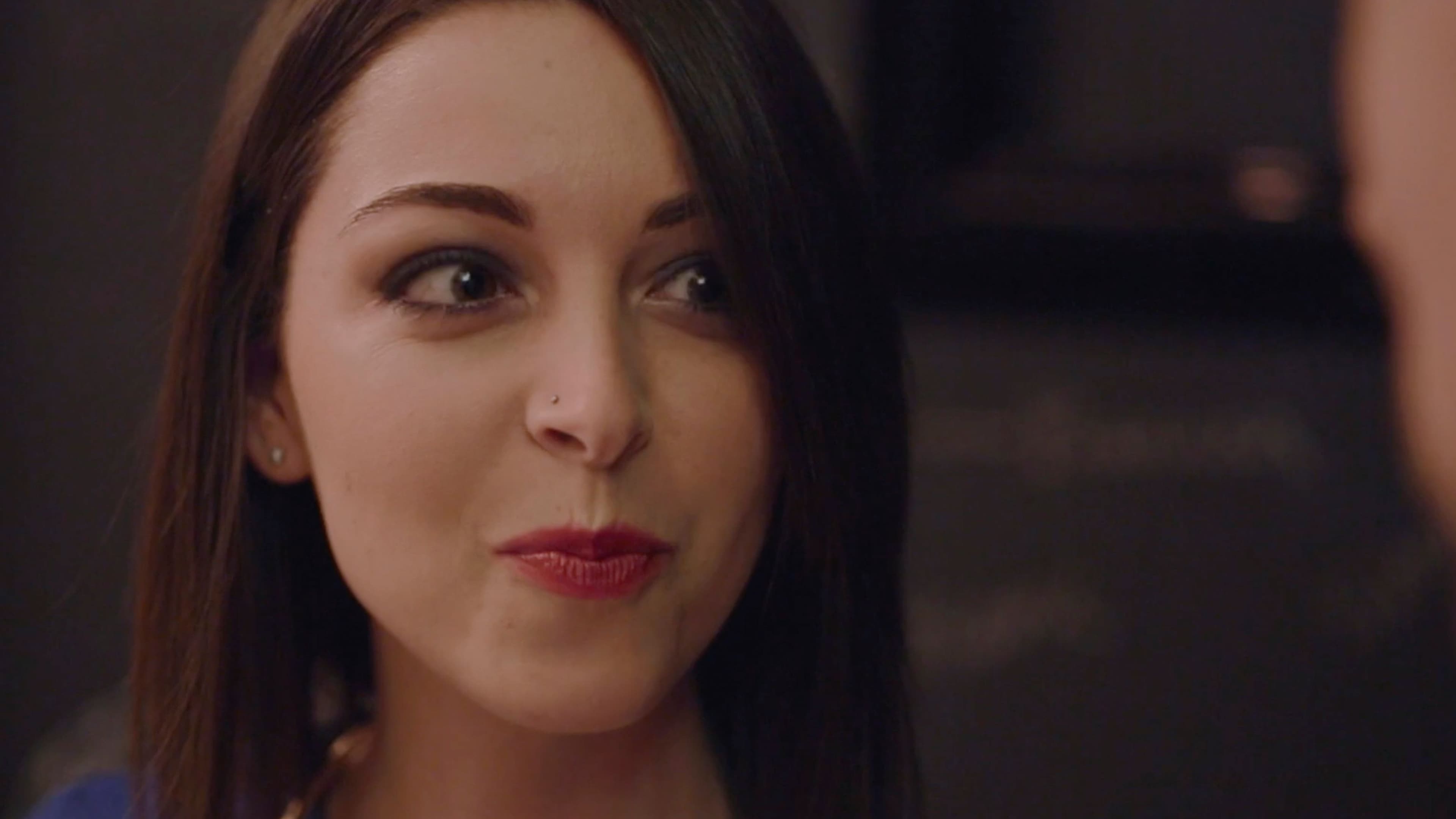 A close-up portrait of a diner with dark hair and red lipstick, glancing sideways with a slight smile in the dimly lit restaurant.