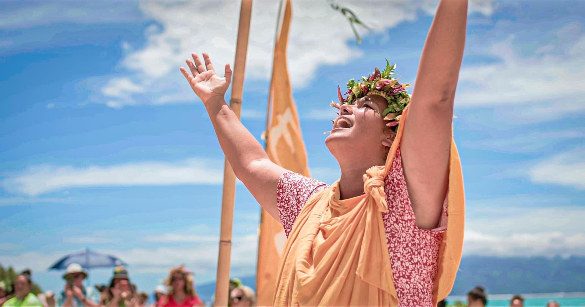 A Tahitian woman wearing a flower crown and orange sash raises her arms skyward mid-chant at a beachside gathering, with a crowd and turquoise lagoon behind her.