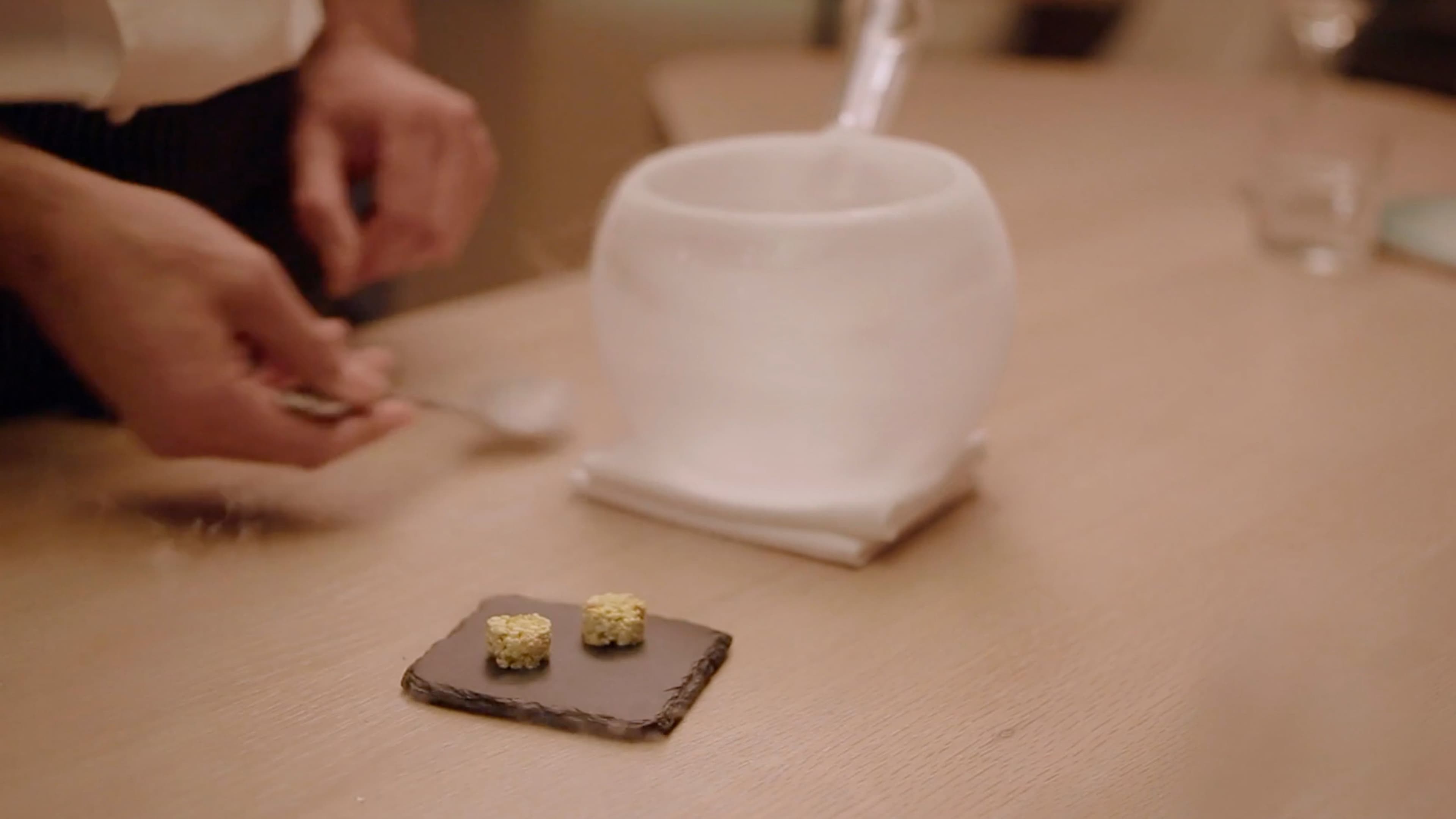 A chef's hands carefully plate two small golden bites onto a slate tile beside a white ceramic vessel at the counter.