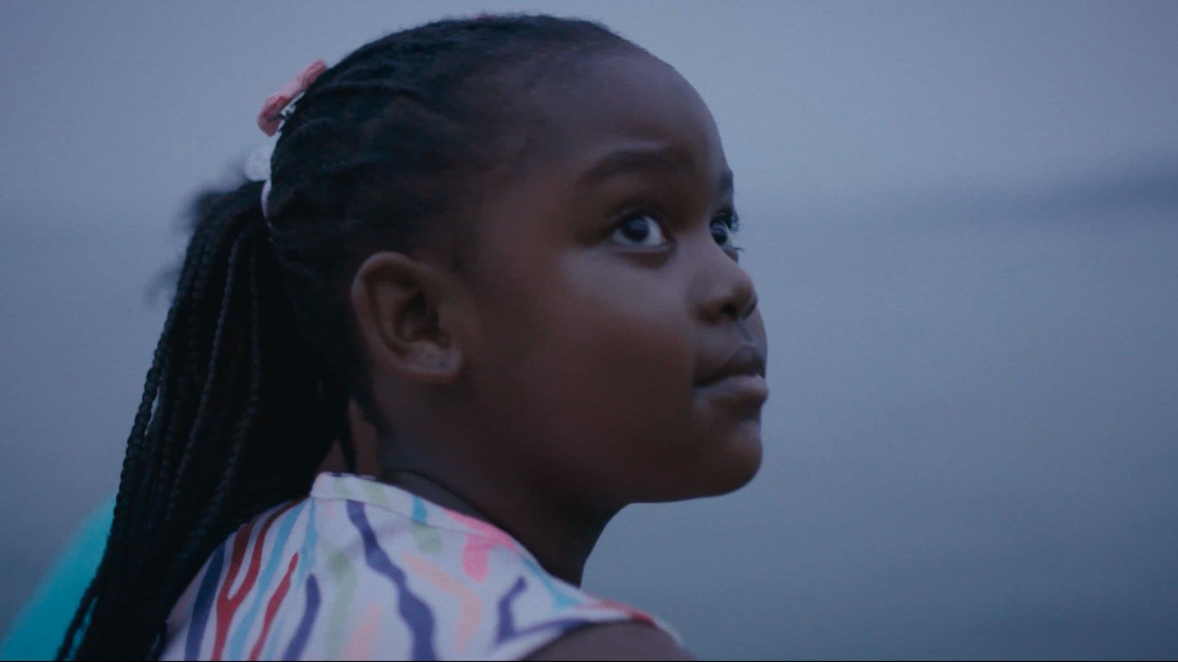 A close-up profile of a young Black girl with braided hair and a colourful patterned top, gazing upward against a soft grey sky. The intimate cinematic framing conveys quiet contemplation and hope.
