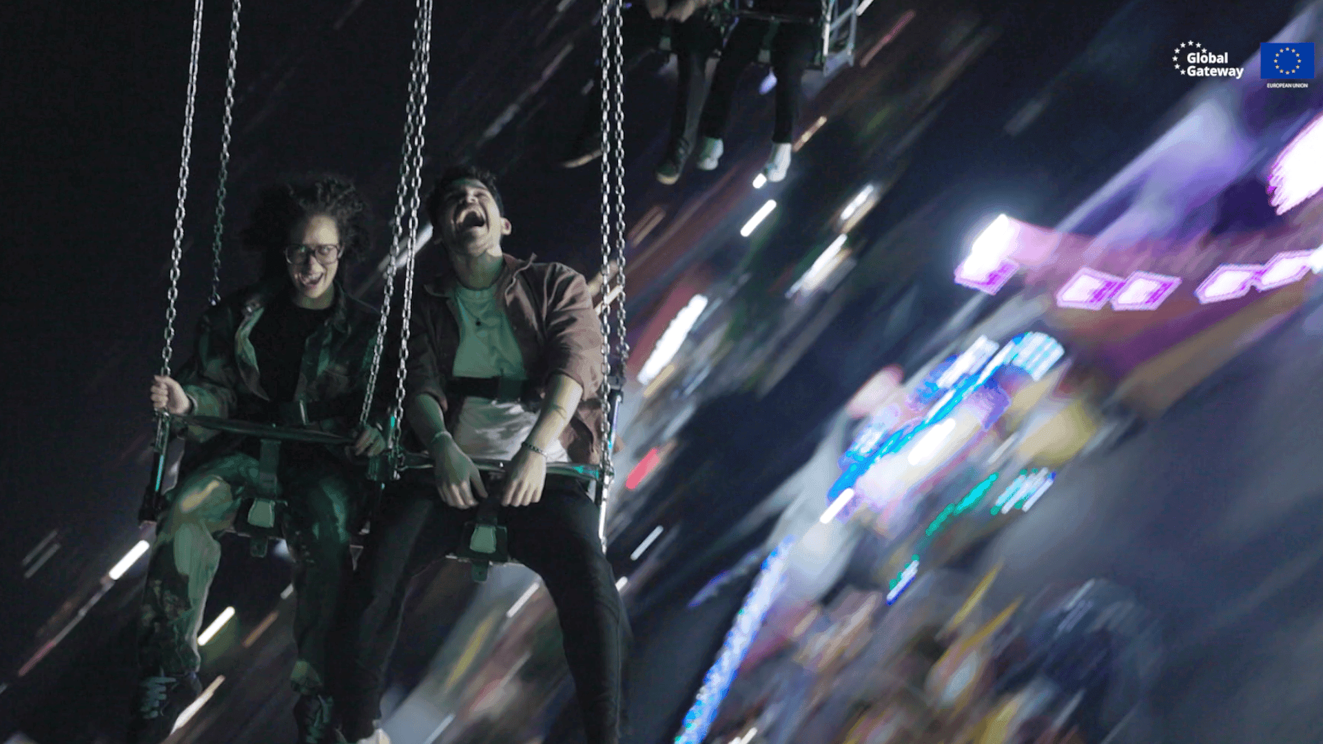 Two people laugh and scream joyfully on a swing carousel ride at night, colourful lights streaking past in motion blur.