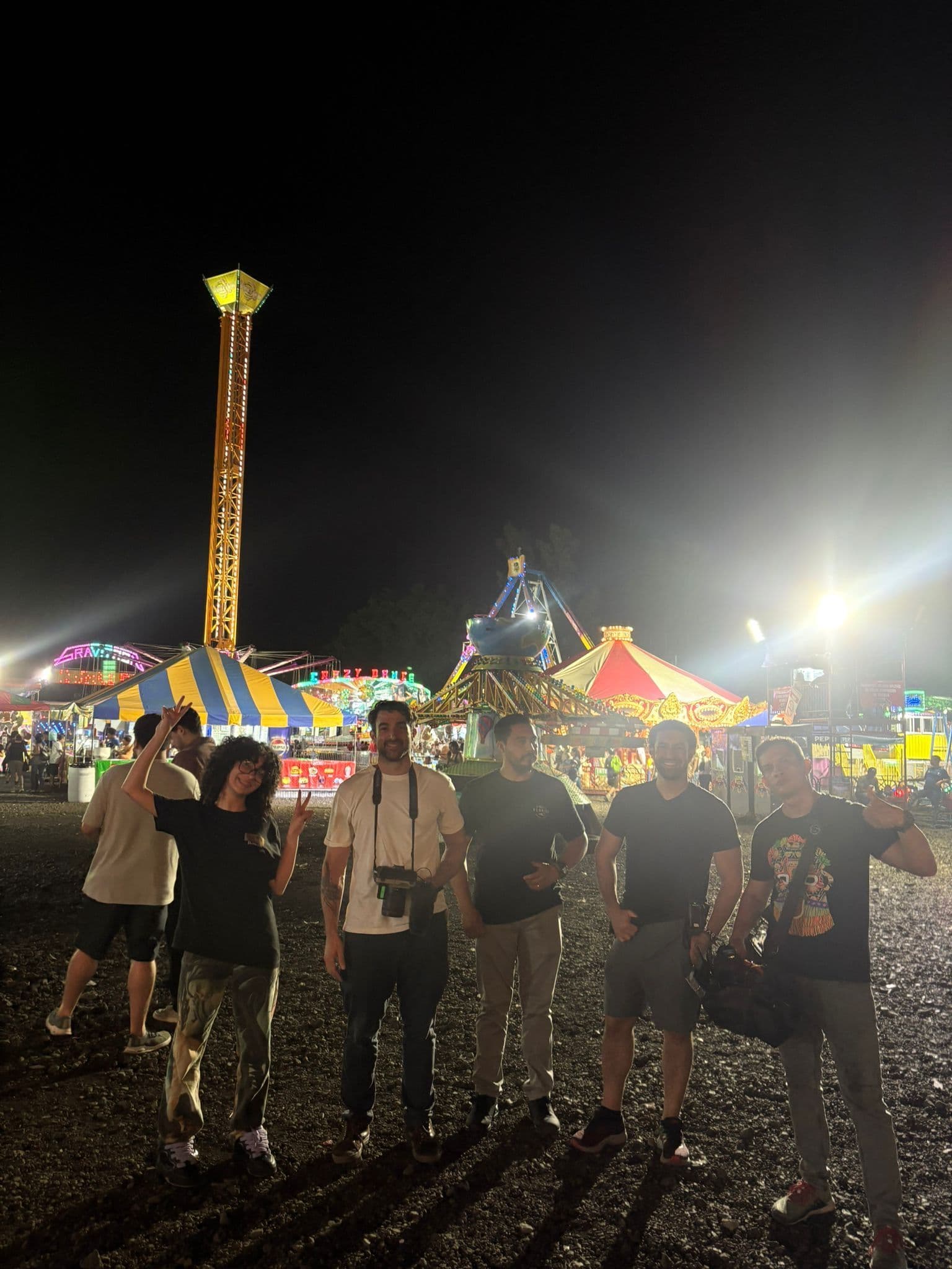 A small group from the crew poses together at a colourful carnival at night, fairground rides lit up behind them.