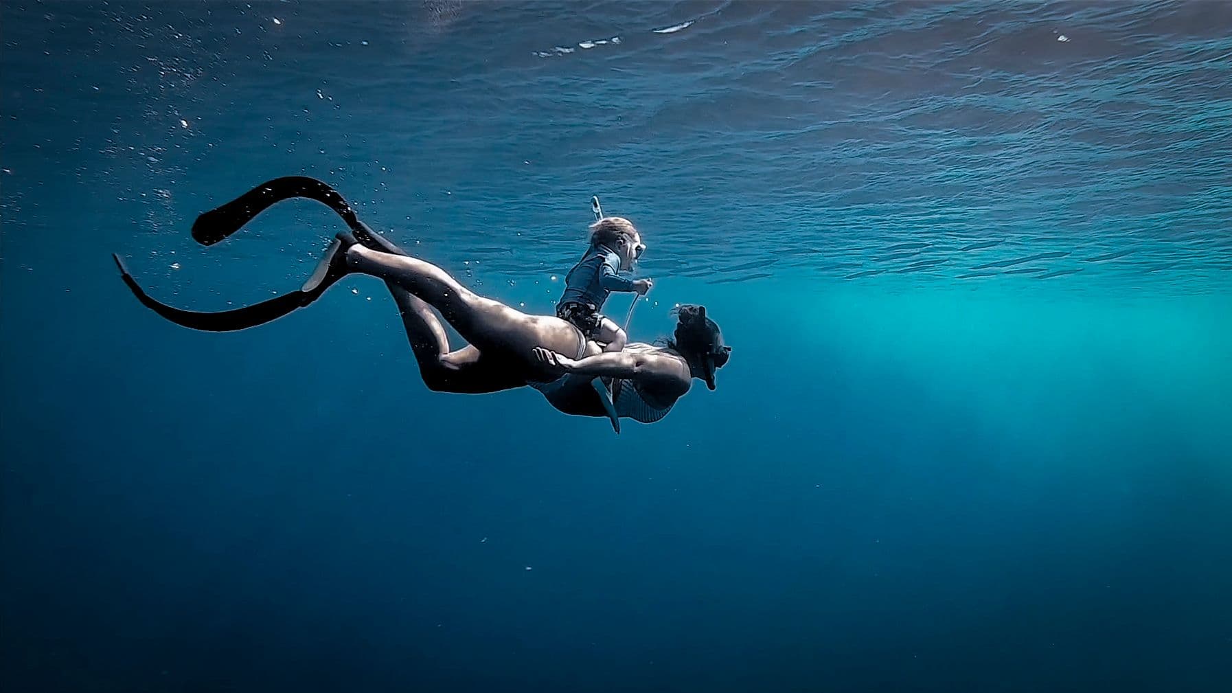 A freediver in mask and fins descends gracefully through deep blue water, sunlight filtering down from the surface above.