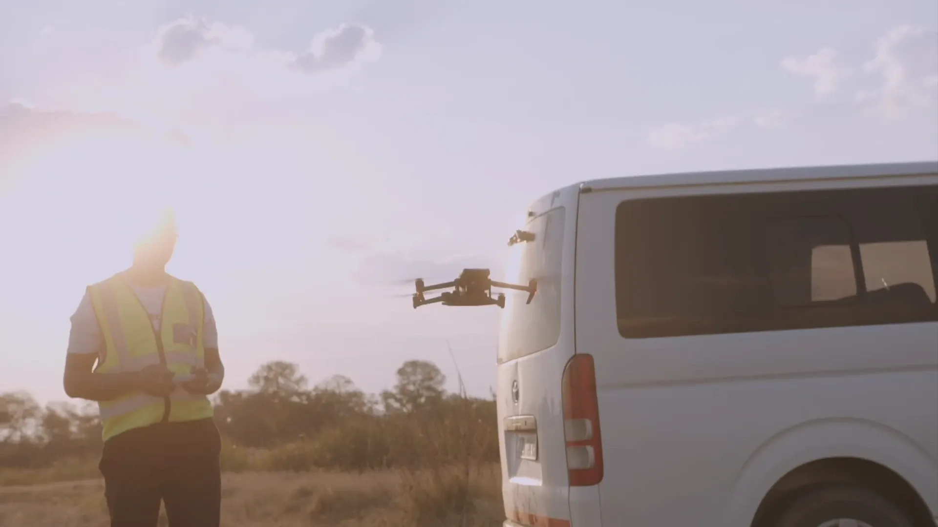 A drone operator in a high-visibility vest flies a drone near a parked van in a field.