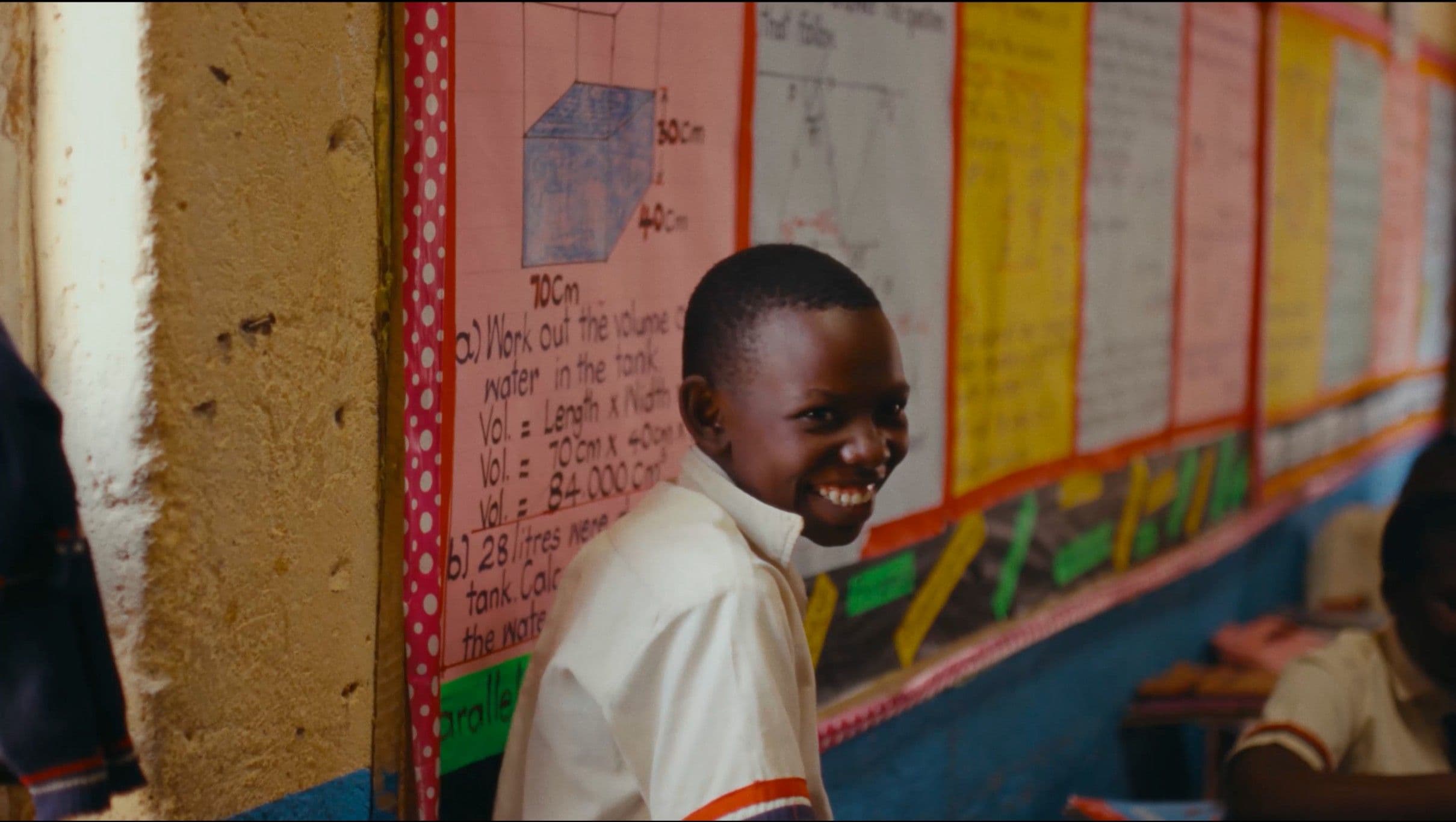 A young Black schoolboy in a white uniform turns to flash a wide, joyful smile at the camera inside a colourful classroom. Educational posters and maths charts line the walls behind him.