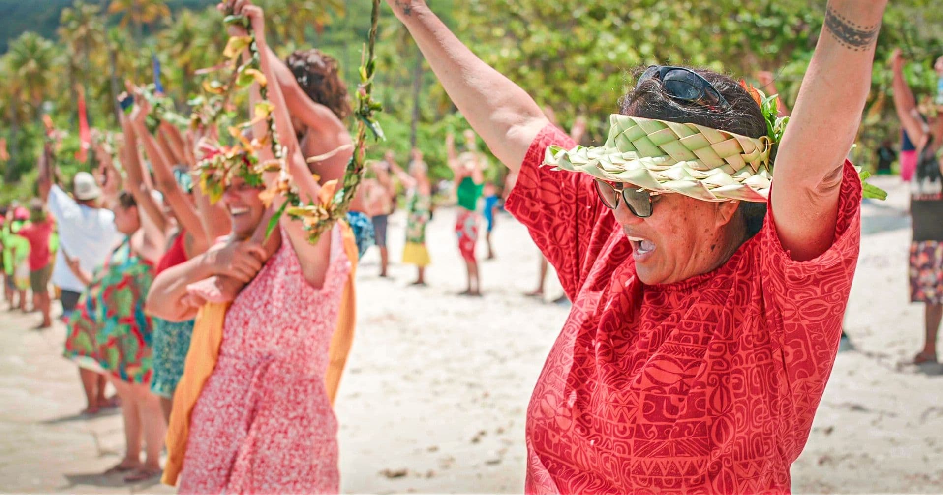 A line of community members in colourful patterned clothing and woven palm-leaf hats raise their arms joyfully together on the beach.