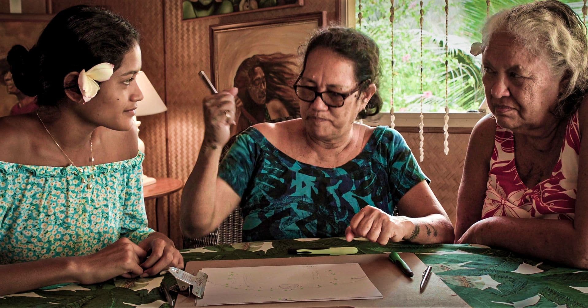 Three women sit at an indoor table reviewing handwritten notes together, deep in conversation.