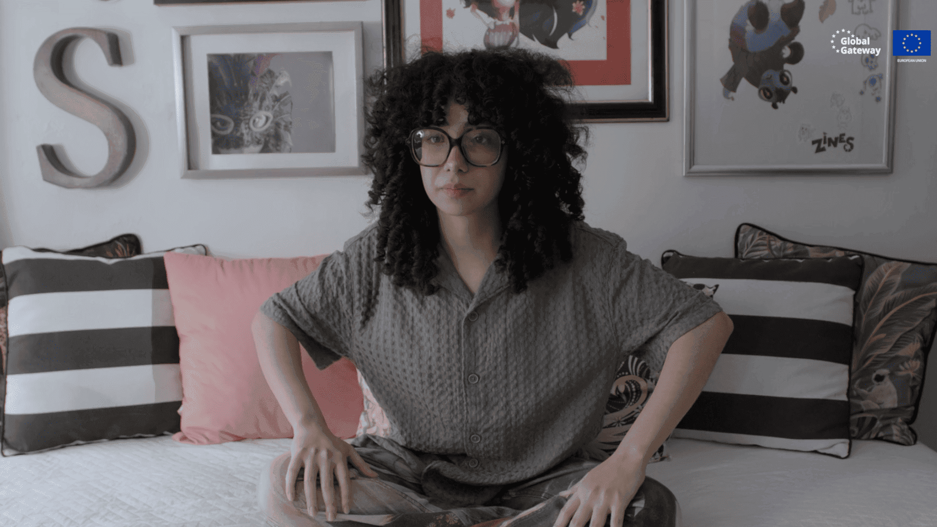 Valeria sits cross-legged on a bed in her personal room, decorated with striped and pink pillows and framed artwork, looking directly at the camera.