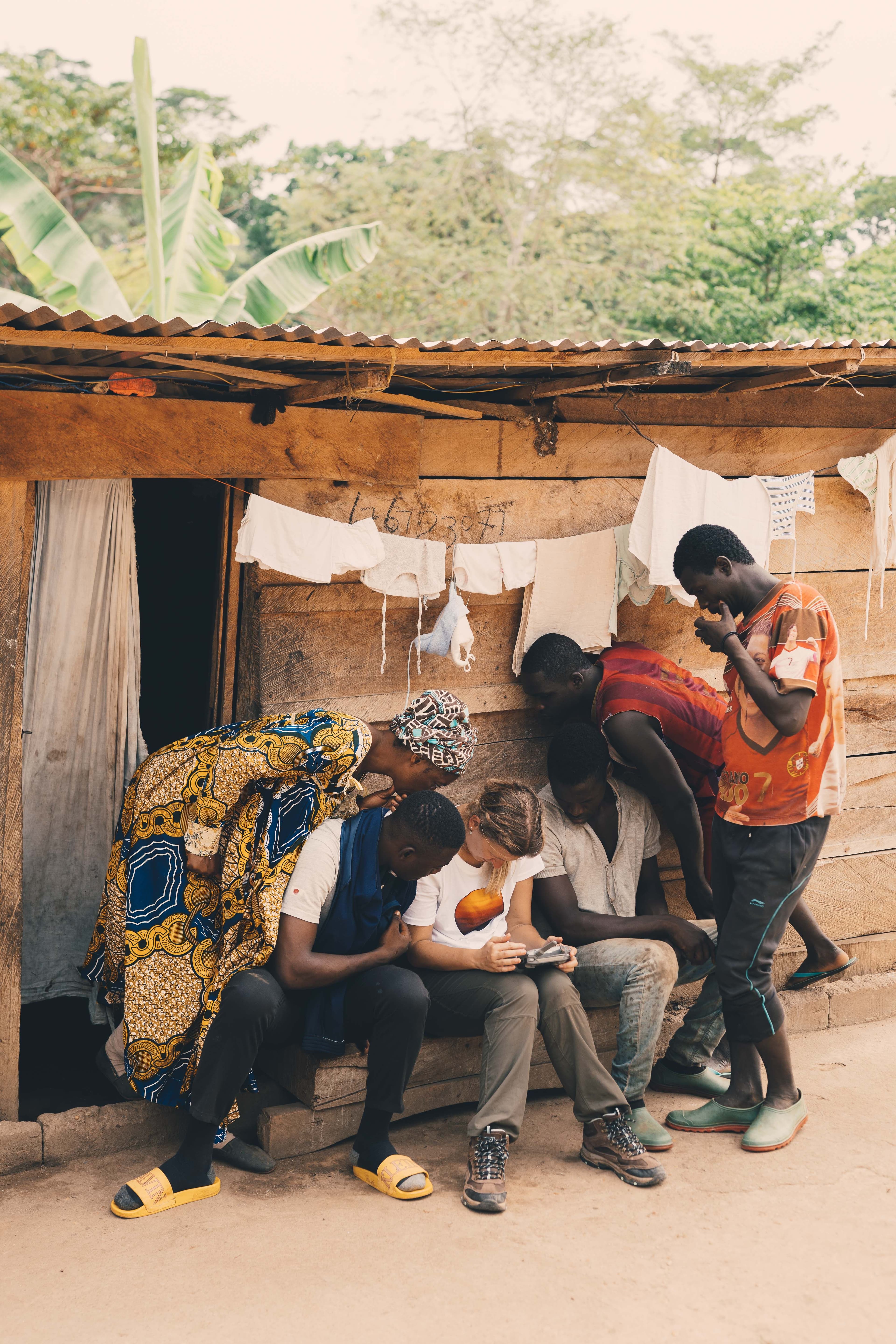 A group of local residents and crew members gather around a handheld screen outside a wooden house, reviewing footage together.