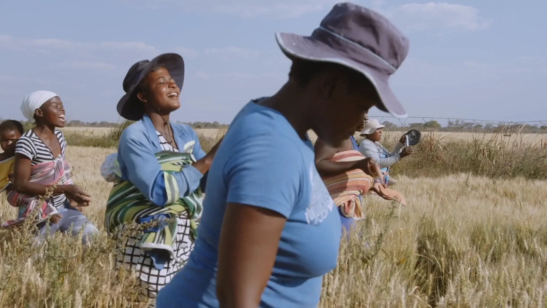 A group of women laugh together while working in a field, wearing sun hats.