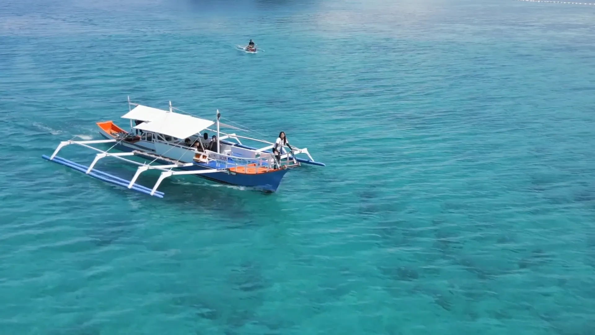 An aerial view of a traditional outrigger boat on clear turquoise water, with a person on a paddleboard nearby.