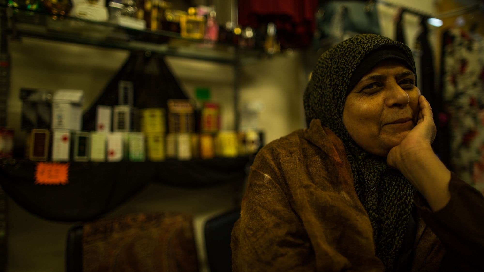 An older woman in a dark patterned headscarf and brown shawl rests her cheek on her hand and smiles softly, seated in a shop lined with shelves of perfume boxes.